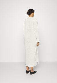 Femme aux cheveux bouclés foncés, portant une longue robe blanche en lin et des chaussures noires sans lacets, debout de dos sur un fond blanc uni.