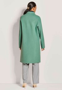 Lichtgroene, lange jas met een gestructureerde kraag, gladde textuur en rechte snit. Draagt over grijze broek en beige hakken.