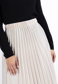 Plisseret cremefarvet nederdel med elastisk talje. Stoffet ser glat ud med en flydende tekstur, som bliver komplementeret af en sort bluse med lange ærmer.