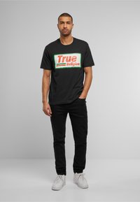 Tricou din bumbac negru, cu un design îndrăzneț al logo-ului "True Religion" în roșu și verde, asociat cu pantaloni negri și adidași albi.