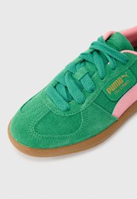 Grønn semsket sneaker med rosa aksent, med teksturerte snorer og såle av gummi. Gullmerke på siden gir en bemerkelsesverdig detalj.
