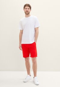 Camiseta blanca de algodón combinada con pantalones cortos rojos de algodón, con dobladillos enrollados, y complementada con zapatillas blancas sobre un fondo neutro.