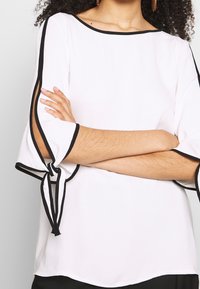 Blouse blanche avec des bords noirs, présentant des manches cloche avec des liens au niveau des coudes. Texture lisse, encolure ronde et coupe décontractée.