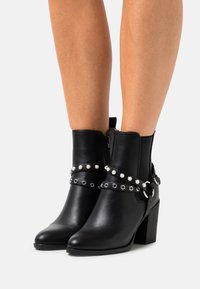 Botines de cuero negro hasta el tobillo con tacón cuadrado, que presentan tachuelas plateadas y correas decorativas. Cierre con cremallera en el lateral. Textura suave.