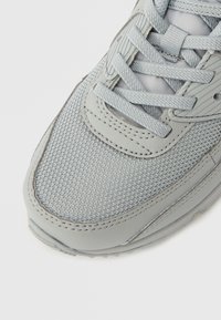 Szürke sportcipő texturált hálós felsőrésszel, sima bőrrészletekkel, lapos fűzőkkel és párnázott talppal. A közeli nézet kiemeli a részleteket.