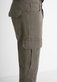 Pantaloni cargo marroni con molte tasche a patta e cuciture visibili, mostrati di lato su uno sfondo neutro.