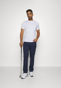 Witte T-shirt met korte mouwen en logo, marineblauwe broek en witte sportschoenen. De persoon houdt een golfclub vast en staat tegen een effen achtergrond.