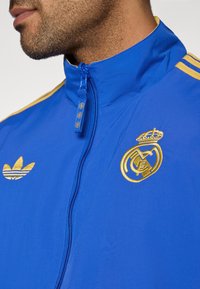 Blue jacket made of lightweight material with gold accents. Features a high collar, zip closure, and embroidered logos of Adidas and Real Madrid.