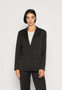 Black tailored blazer with a single button closure, sharp lapels, two front pockets, and a smooth fabric texture, paired with a light top.