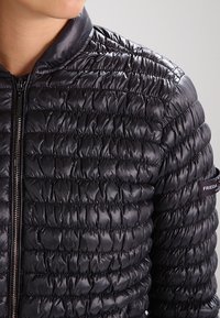 Zwarte pufferjack in een geborduurd ontwerp met een glanzende, soepele textuur. Voorzien van een volledige rits en een merkaccent op de mouw.