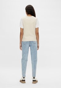 Femme debout de dos portant un pull sans manches en tricot crème par-dessus un t-shirt blanc, un jean bleu clair et des chaussures beiges épaisses.