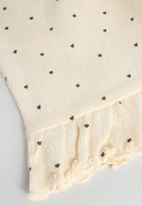 Tela color crema con pequeños patrones de corazones negros. Presenta un dobladillo con volantes en la parte inferior, añadiendo textura al diseño.