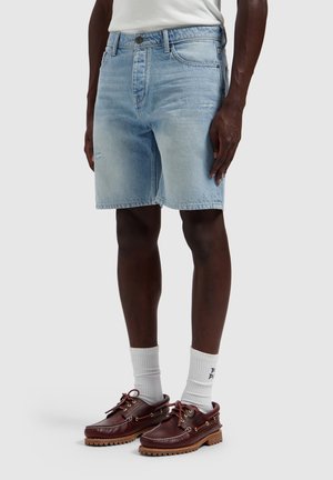 Man draagt lichtblauwe spijkershorts, witte sokken en bruine leren bootschoenen, staand tegen een effen achtergrond.