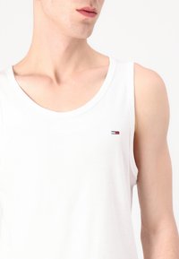 Νέος άνδρας που φοράει λευκό αμάνικο τοπ με μικρό κόκκινο, λευκό και μπλε λογότυπο στο αριστερό στήθος, εμφανίζεται από τους ώμους μέχρι το πηγούνι.