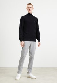 Zwarte gebreide trui met hoge kraag en rits, gecombineerd met lichtgrijze op maat gemaakte broek en witte sneakers. Eenvoudig, gestroomlijnd ontwerp.