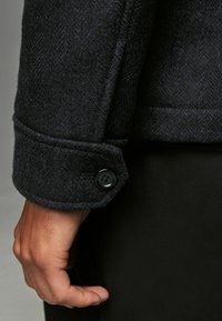 Dunkelblauer Wollmantel mit Fischgrätenmuster, ausgestattet mit einem geknöpften Ärmelabschluss und schwarzen Knopfdetails sowie einer glatten Textur.