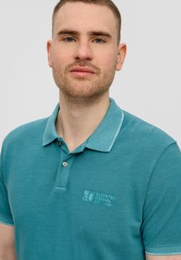 Tyrkysová polo košile s texturovaným povrchem, kontrastním límečkem a předním logem. Obsahuje dva knoflíky a kratké rukávy.