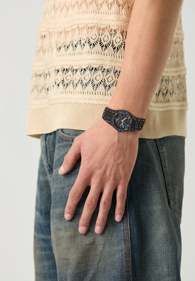 Sort ur med farverige sprøjt, firkantet urskive, gummirem. Båret på en hånd, der hviler på denimstof, med en tekstureret sweater ovenover.