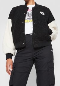 Svart och vit sherpa bomberjacka med ribbad krage, tryckknappar och broderad patch; bärs över en grafisk t-shirt och cargobyxor.