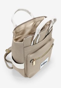 Mochila de lona beige con correas trenzadas blancas, detalles en cremallera dorada, bolsillo frontal y un parche de tela con el logo. Parte superior abierta para un fácil acceso.