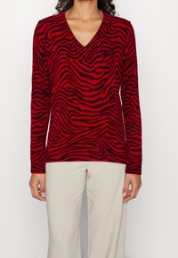 Pull à manches longues rayé rouge et noir avec un col en V, doté d'une texture douce et d'une coupe décontractée, associé à un pantalon de couleur claire.