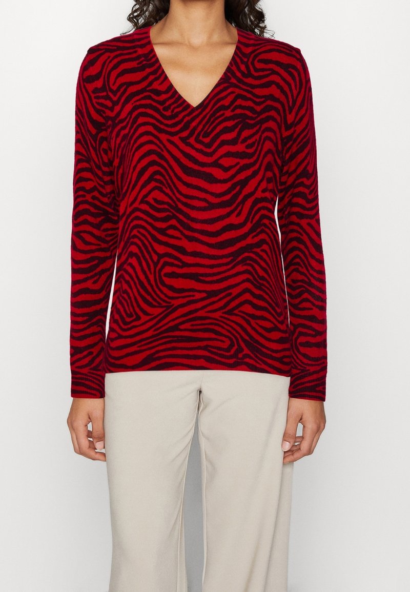 Pull à manches longues rayé rouge et noir avec un col en V, doté d'une texture douce et d'une coupe décontractée, associé à un pantalon de couleur claire.