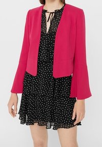 Veste courte fuchsia à manches évasées portée par-dessus une robe noire à pois avec un ourlet volanté et un col noué.