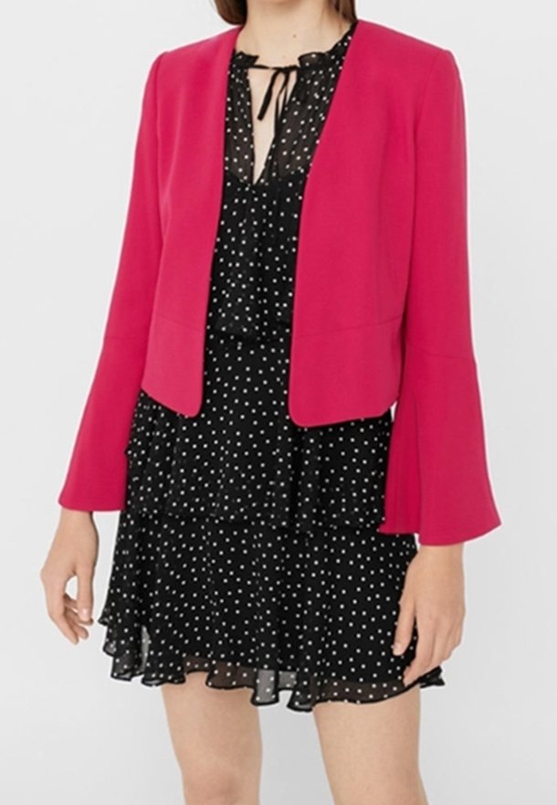 Veste courte fuchsia à manches évasées portée par-dessus une robe noire à pois avec un ourlet volanté et un col noué.