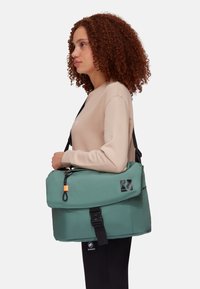 Borsa a tracolla verde in tessuto con chiusura a patta, dettagli neri, tracolla regolabile e patch con logo. Texture liscia con una finitura resistente.