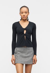 Γυναίκα με πλεκτά μαλλιά που φοράει μαύρο μακρυμάνικο τοπ με κεντρική στριφτή λεπτομέρεια και ασπρόμαυρη καρό μίνι φούστα.