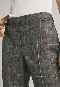 Personne ajustant un pantalon à carreaux gris avec des lignes rouges et blanches, portant un haut côtelé blanc, coupé à la taille.