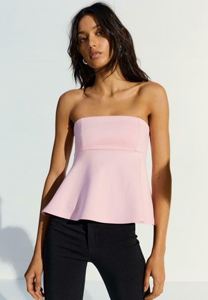 Žena s dugom tamnom kosom koja nosi svijetloružičasti peplum top bez naramenica i crne uske hlače, stoji na jednostavnoj svijetloj pozadini.