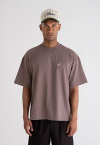 Brown oversized t-shirt with a round neckline and short sleeves, featuring a small white logo on the chest; worn with black shorts.