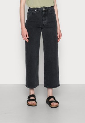 Person iført sorte vidtstrakte jeans med høj talje og sorte sandaler med dobbeltremme mod en ensfarvet lys baggrund.