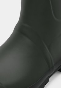 Botte en caoutchouc verte avec une texture lisse, forme arrondie et légères plis près de la cheville ; dotée d'une semelle noire durable pour une meilleure adhérence.