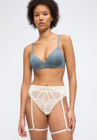 Γυναίκα με κοντά σγουρά μαλλιά φορά γκρι σουτιέν και λευκή δαντελένια ζώνη για καλτσοδέτες με καλσόν, σε φόντο απλό και φωτεινό.