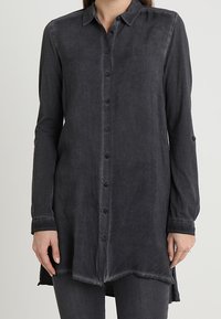Donna che indossa una camicia lunga grigio scuro con bottoni, colletto e maniche lunghe, abbinata a pantaloni grigio scuro.