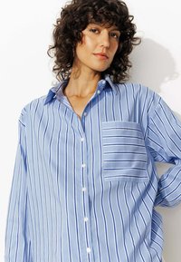 Chemise bleu clair à manches longues avec des rayures verticales blanches et marines, dotée d'une poche poitrine et d'une coupe décontractée, fabriquée en tissu lisse.