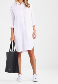 Biała sukienka koszulowa z trzema ćwierć rękawami, bocznymi rozcięciami i luźnym krojem. W zestawie z czarną torbą typu tote i białymi butami wsuwanymi.