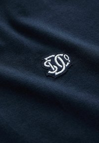 Mörkblått bomullstyg med en slät textur, med en vit broderad logotypdetalj i nedersta hörnet.