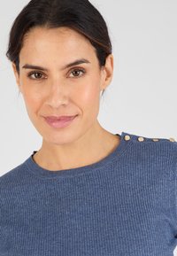 Sweater bleu à côtes avec un col rond et des boutons décoratifs dorés sur l'épaule. Gros plan sur la texture et la couleur du tissu.