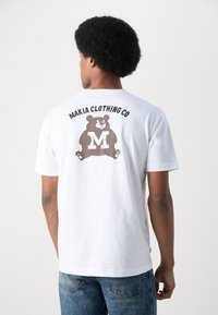 Bílý bavlněný tričko s potiskem hnědého medvěda a textem "MAKIA CLOTHING CO" nad ním. Krátké rukávy a standardní střih.