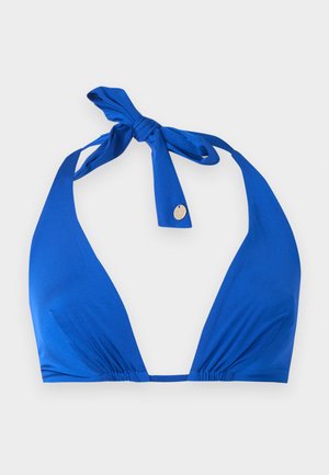 Femeie stând pe un fundal alb, purtând un top de bikini albastru cu după gât, slipuri negre cu talie înaltă și sandale negre, cu o mână pe șold.