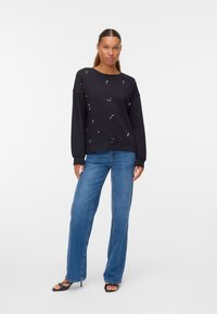 Sudadera negra con tachuelas blancas dispersas, cuello redondo y mangas largas. Combinada con jeans acampanados azul claro y sandalias de tacón negras.