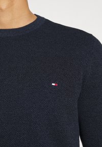 Granatowy sweter dzianinowy z okrągłym dekoltem i prążkowaną teksturą, z małym czerwono-białe-niebieskim logo na piersi.