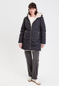 Abrigo negro acolchado con capucha forrada de piel, cremallera frontal y bolsillos laterales. Combinado con jeans grises y zapatillas blancas.