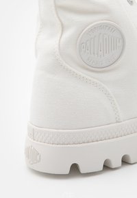 Chaussure montante en toile blanche Palladium avec une semelle en caoutchouc texturée et un patch logo circulaire sur le côté près de la cheville.
