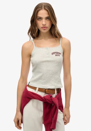 Jonge vrouw met golvend haar draagt een lichtgrijze geribbelde tanktop met de tekst "Superdry 1979", witte broeken, een bruine riem en een vastgebonden rode sweatshirt.