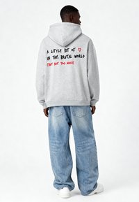 Pelēka kapuce ar aizmugures tekstu: "Mazliet ❤️ šajā brutālajā pasaulē (bet ne pārāk daudz)," apvienota ar gaiši ziliem platajiem džinsiem un baltām kedām.