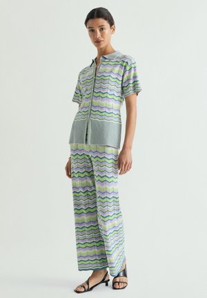 Mujer con camisa de manga corta y patrón de zigzag en tonos pastel a juego y pantalones de pierna ancha con tacones abiertos negros, de pie sobre fondo blanco.